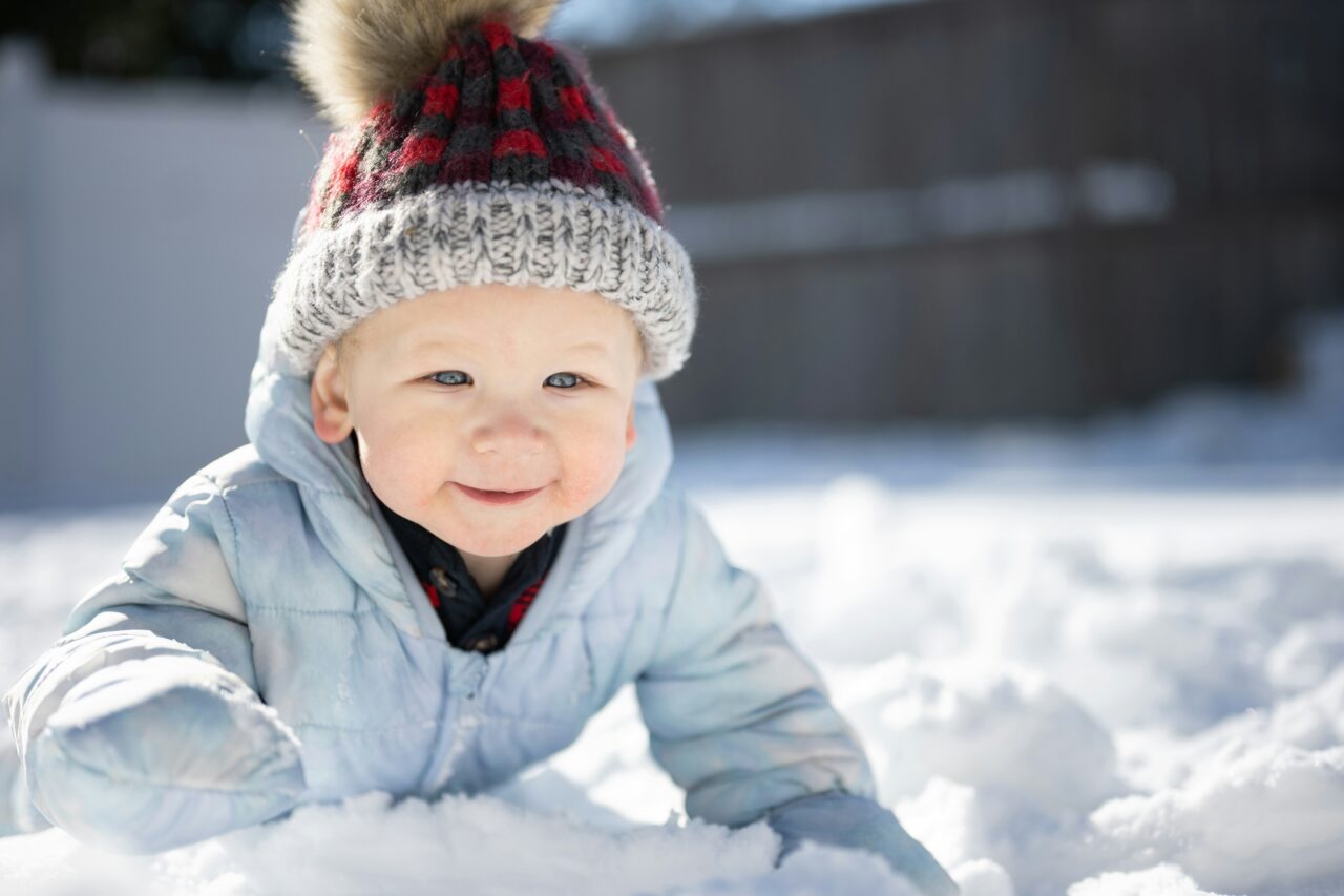 baby in a red hat and blue jacket on stomach in the snow